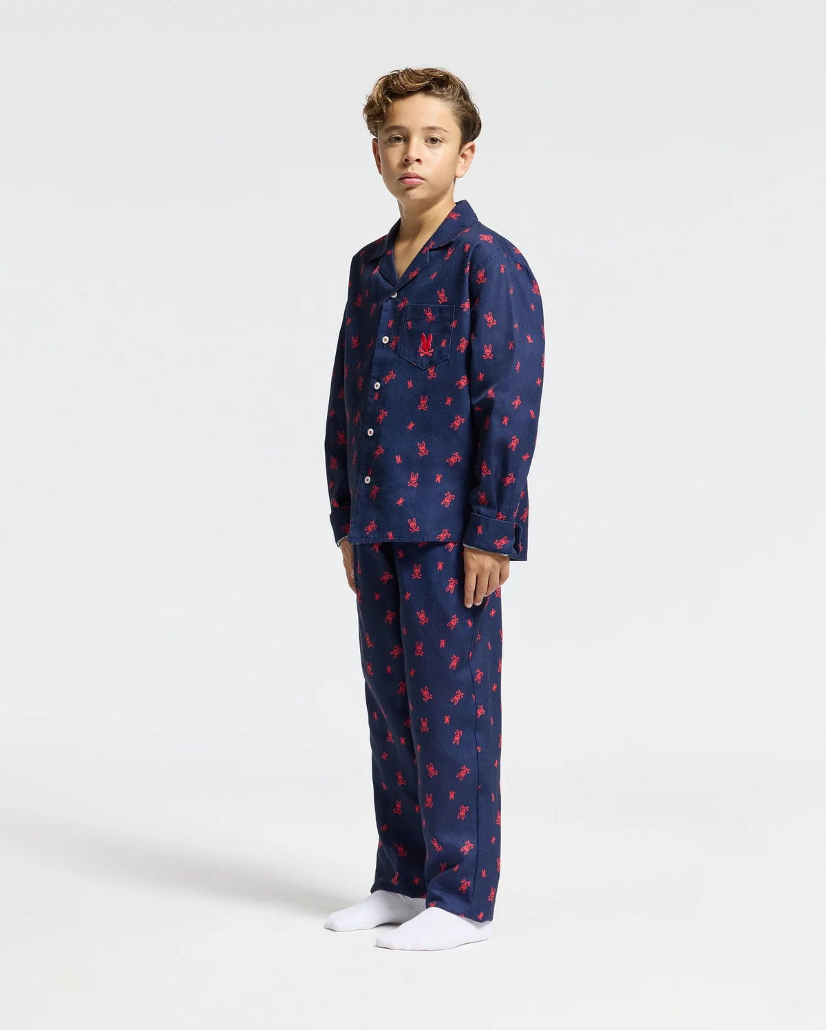 A young boy stands against a white background, dressed in a Psycho Bunny KIDS ALL OVER PRINT FLANNEL PAJAMA SET (B0X356E200), featuring dark blue pajamas adorned with a red crab pattern. His collared shirt and matching pants are perfectly complemented by his white socks and short, curly hair.