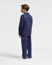 A person is wearing the Psycho Bunny KIDS ALL OVER PRINT FLANNEL PAJAMA SET - B0X356E200 in navy blue with a red pattern, standing facing away and looking to the left. The background is plain white, and they are wearing white socks.