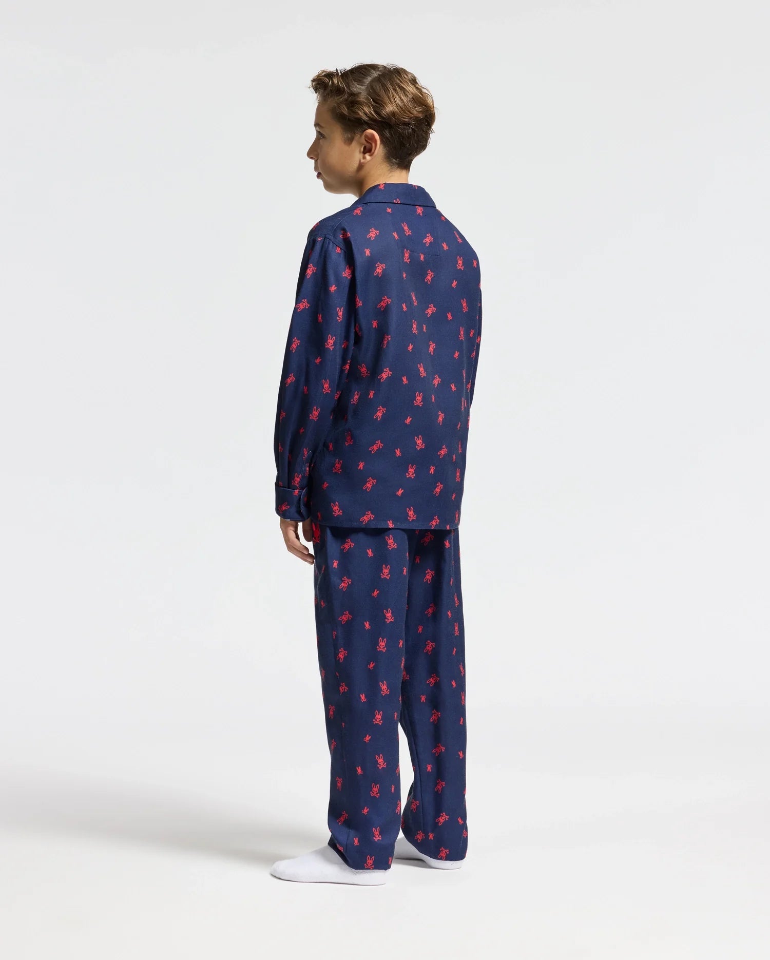 A person is wearing the Psycho Bunny KIDS ALL OVER PRINT FLANNEL PAJAMA SET - B0X356E200 in navy blue with a red pattern, standing facing away and looking to the left. The background is plain white, and they are wearing white socks.