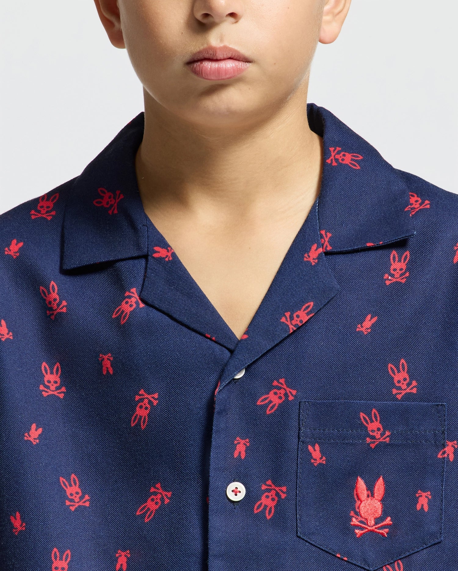 A person wearing the Psycho Bunny KIDS ALL OVER PRINT FLANNEL PAJAMA SET in navy blue. This shirt, designed like a flannel sleep set for children, is adorned with small red outlines of bunnies across the fabric and includes a pocket on the left side. The bunnies are arranged in varying orientations throughout the design.