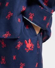 Close-up of the KIDS ALL OVER PRINT FLANNEL PAJAMA SET by Psycho Bunny, featuring a blue fabric adorned with small red skull and bunny motifs. A hand is partially visible in the pocket, highlighting the playful and contrasting design ideal for cozy nights.