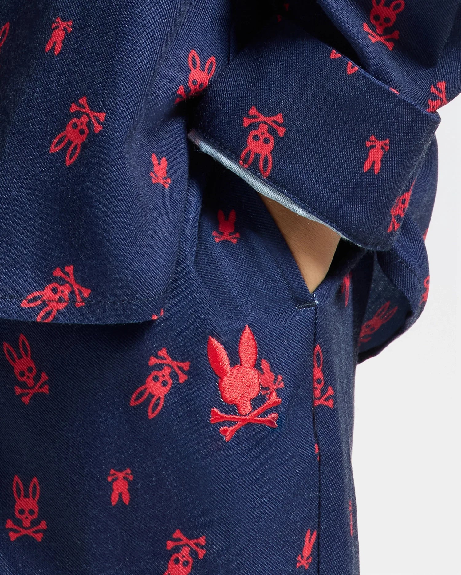 Close-up of the KIDS ALL OVER PRINT FLANNEL PAJAMA SET by Psycho Bunny, featuring a blue fabric adorned with small red skull and bunny motifs. A hand is partially visible in the pocket, highlighting the playful and contrasting design ideal for cozy nights.