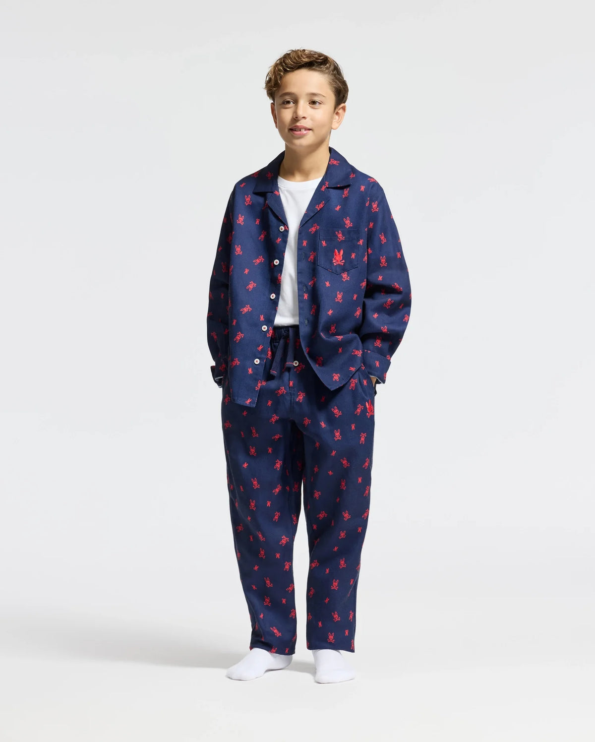 A young boy stands wearing the Psycho Bunny KIDS ALL OVER PRINT FLANNEL PAJAMA SET in navy blue with red patterns. His hands are in his pockets, and he has a relaxed expression. The plain white background accentuates the coziness of this lightweight flannel pajama set for kids.