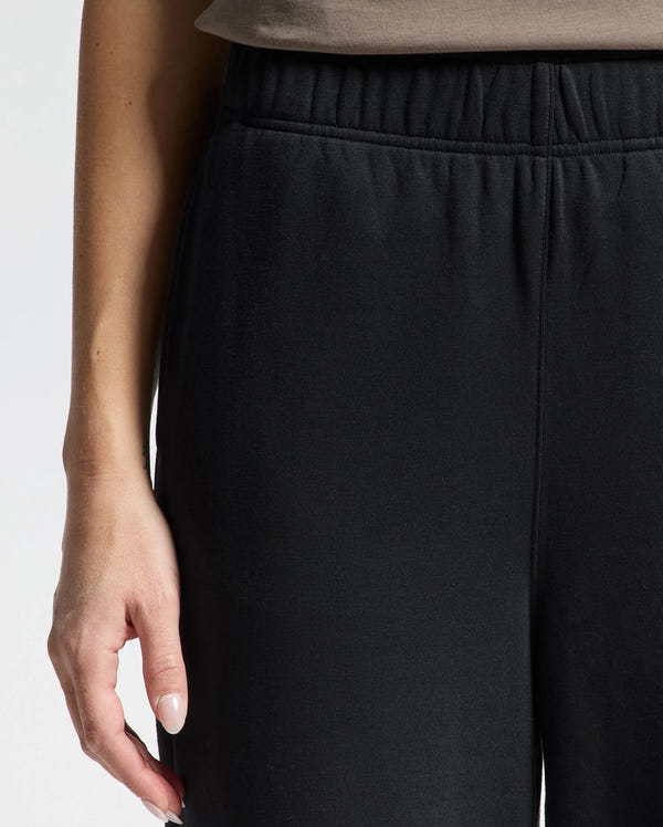 Close-up of a person wearing the Psycho Bunny WOMENS ALEXANDER SWEATPANT - B2P157E200 in black, featuring an elastic waistband. Their right hand rests by their side, accentuating the stylish knit joggers. The plain, light-colored background highlights the sleek design.