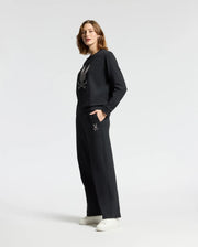 A person models the Psycho Bunny WOMENS ALEXANDER SWEATPANT - B2P157E200 in black featuring a graphic design on the front, set against a white backdrop. They have their hands comfortably in the pockets of these stylish cotton blend sweatpants and are paired with white sneakers.