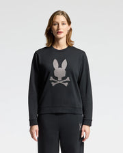 A person stands wearing the WOMENS ALEXANDER SWEATSHIRT - B2S156E200 by Psycho Bunny, featuring a black cotton blend and an embroidered Lurex bunny head above crossed bones. The background is plain white.