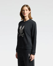 A person with shoulder-length hair is wearing the Psycho Bunny WOMENS ALEXANDER SWEATSHIRT - B2S156E200 in black, featuring an embroidered Lurex bunny skull design. They are paired with matching black pants, set against a plain white background.