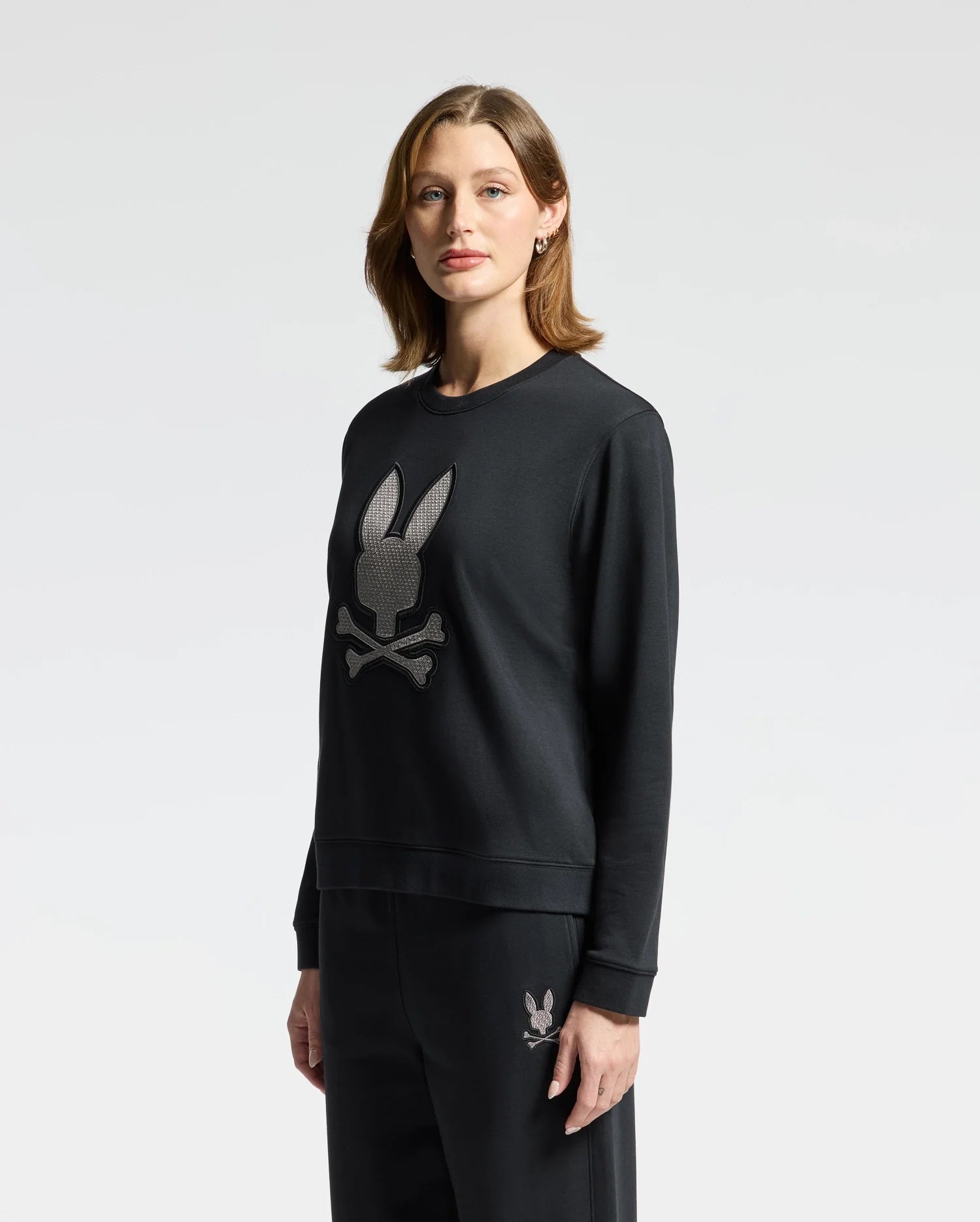 A person with shoulder-length hair is wearing the Psycho Bunny WOMENS ALEXANDER SWEATSHIRT - B2S156E200 in black, featuring an embroidered Lurex bunny skull design. They are paired with matching black pants, set against a plain white background.