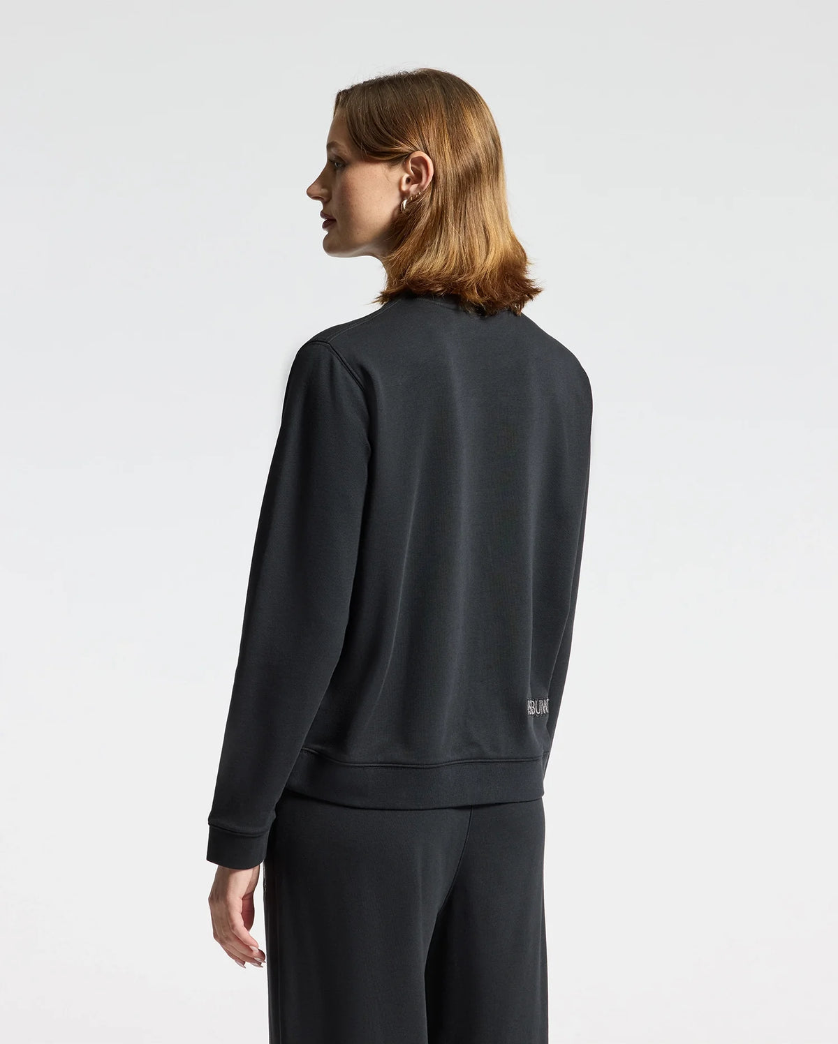 A person with shoulder-length hair stands facing sideways, wearing the WOMENS ALEXANDER SWEATSHIRT - B2S156E200, a black cotton blend from Psycho Bunny featuring an embroidered Lurex bunny, paired with matching pants. The background is plain white.