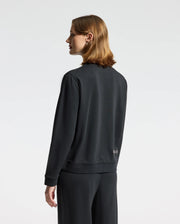 A person with shoulder-length hair stands facing sideways, wearing the WOMENS ALEXANDER SWEATSHIRT - B2S156E200, a black cotton blend from Psycho Bunny featuring an embroidered Lurex bunny, paired with matching pants. The background is plain white.