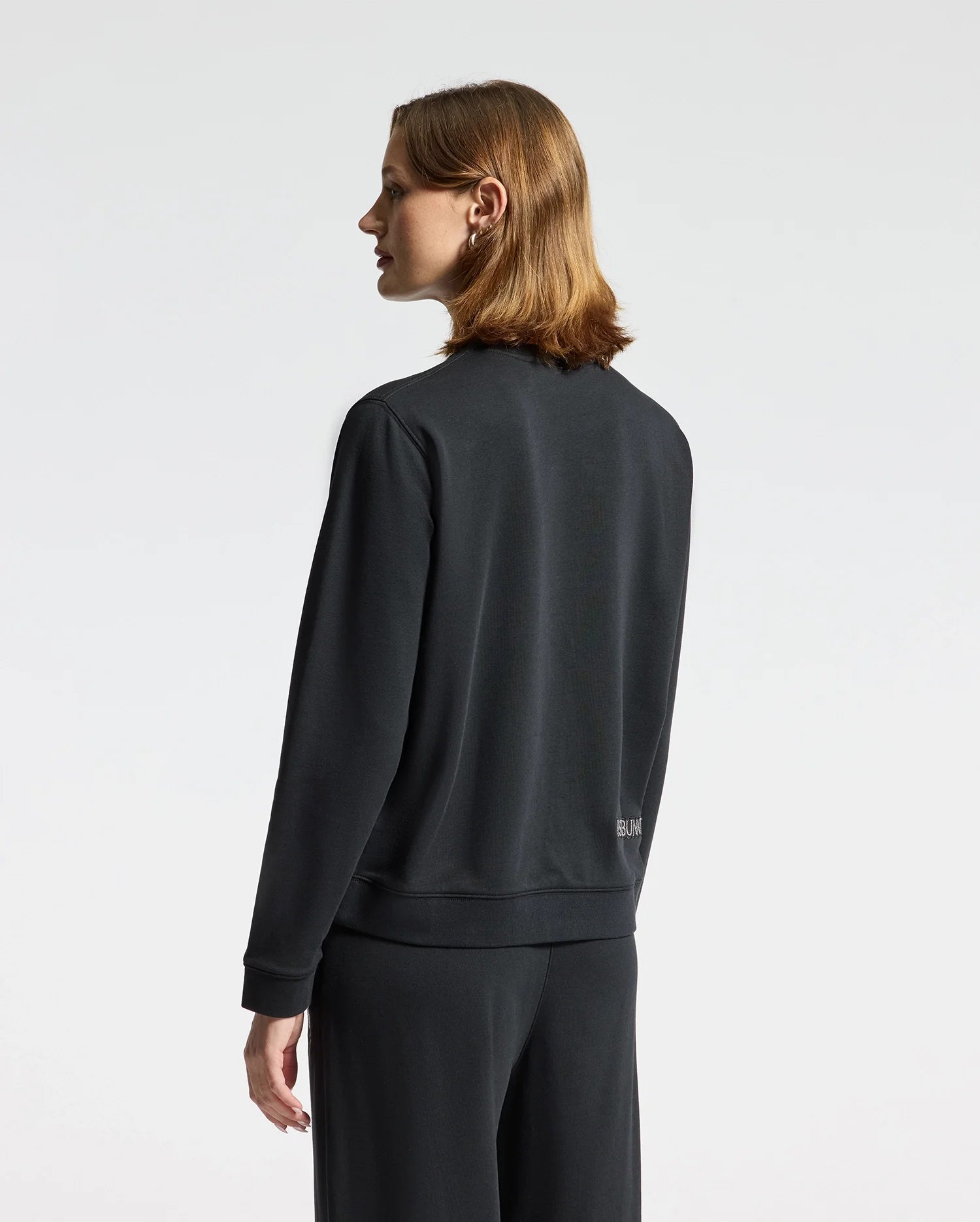 A person with shoulder-length hair stands facing sideways, wearing the WOMENS ALEXANDER SWEATSHIRT - B2S156E200, a black cotton blend from Psycho Bunny featuring an embroidered Lurex bunny, paired with matching pants. The background is plain white.