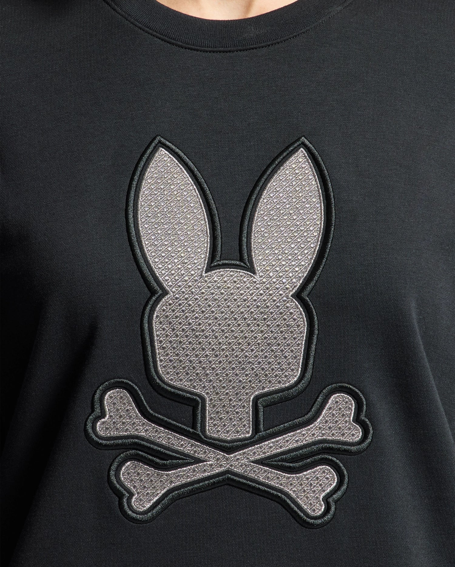 A close-up of the WOMENS ALEXANDER SWEATSHIRT - B2S156E200 by Psycho Bunny highlights an embroidered Lurex bunny design, featuring a stylized animal head with long ears above two crossed bones, evoking a skull and crossbones motif, crafted from a black cotton blend.