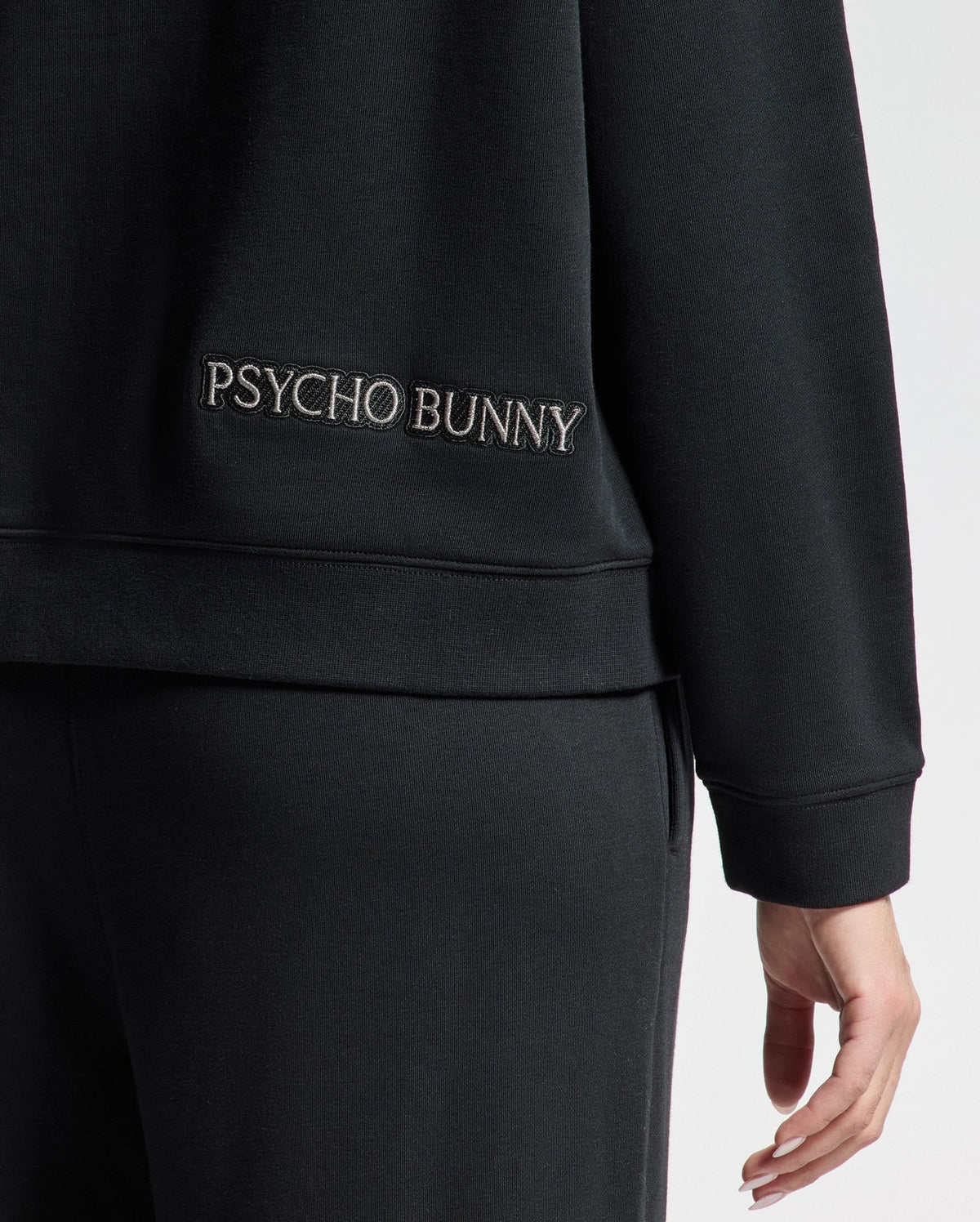 A person is wearing the WOMENS ALEXANDER SWEATSHIRT - B2S156E200 by Psycho Bunny, featuring a black cotton blend design. "PSYCHO BUNNY" is embroidered on the back near the waist, and their hand highlights the intricate Lurex Bunny detail. The emphasis is on this stylish clothing accent.