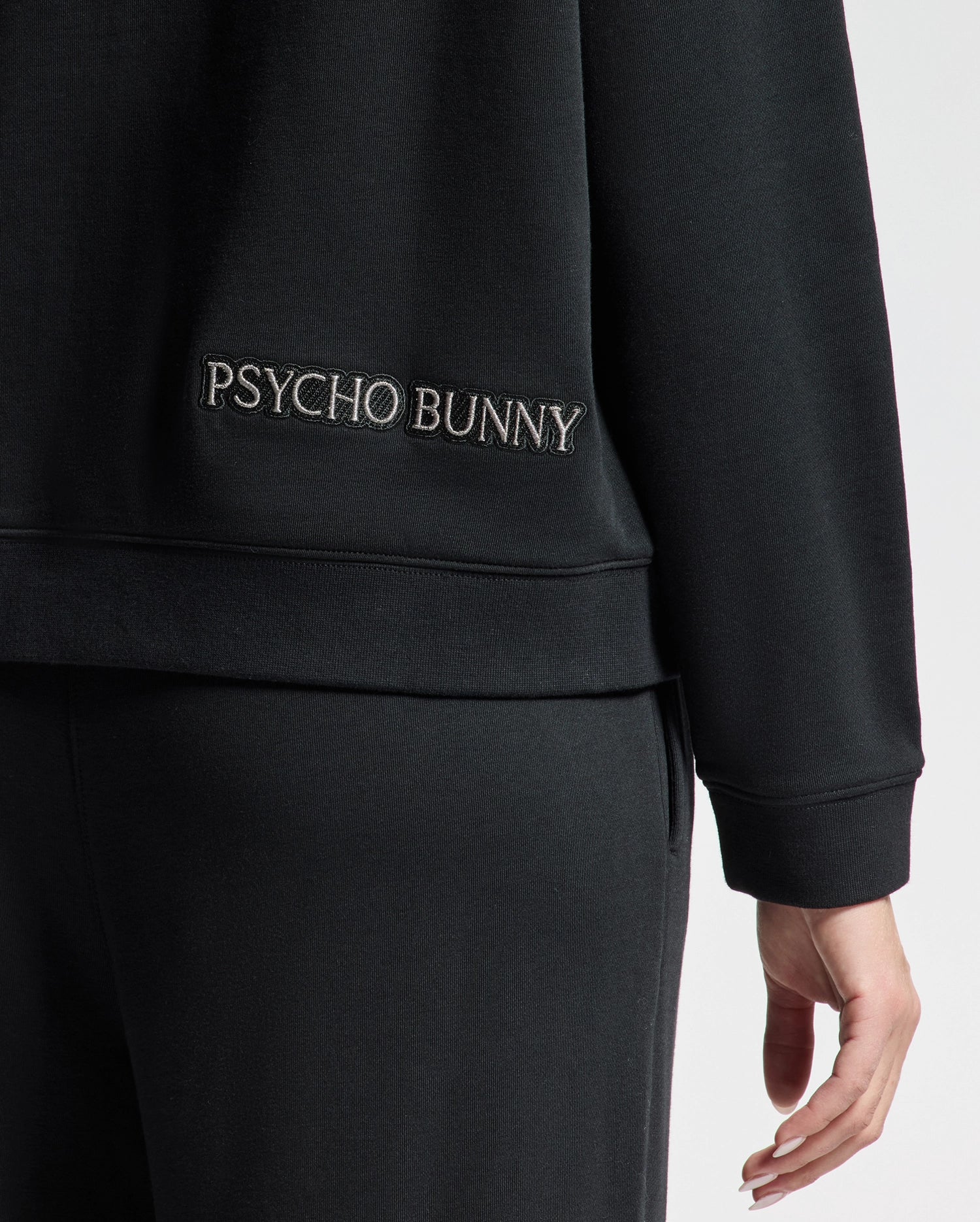 A person is wearing the WOMENS ALEXANDER SWEATSHIRT - B2S156E200 by Psycho Bunny, featuring a black cotton blend design. "PSYCHO BUNNY" is embroidered on the back near the waist, and their hand highlights the intricate Lurex Bunny detail. The emphasis is on this stylish clothing accent.