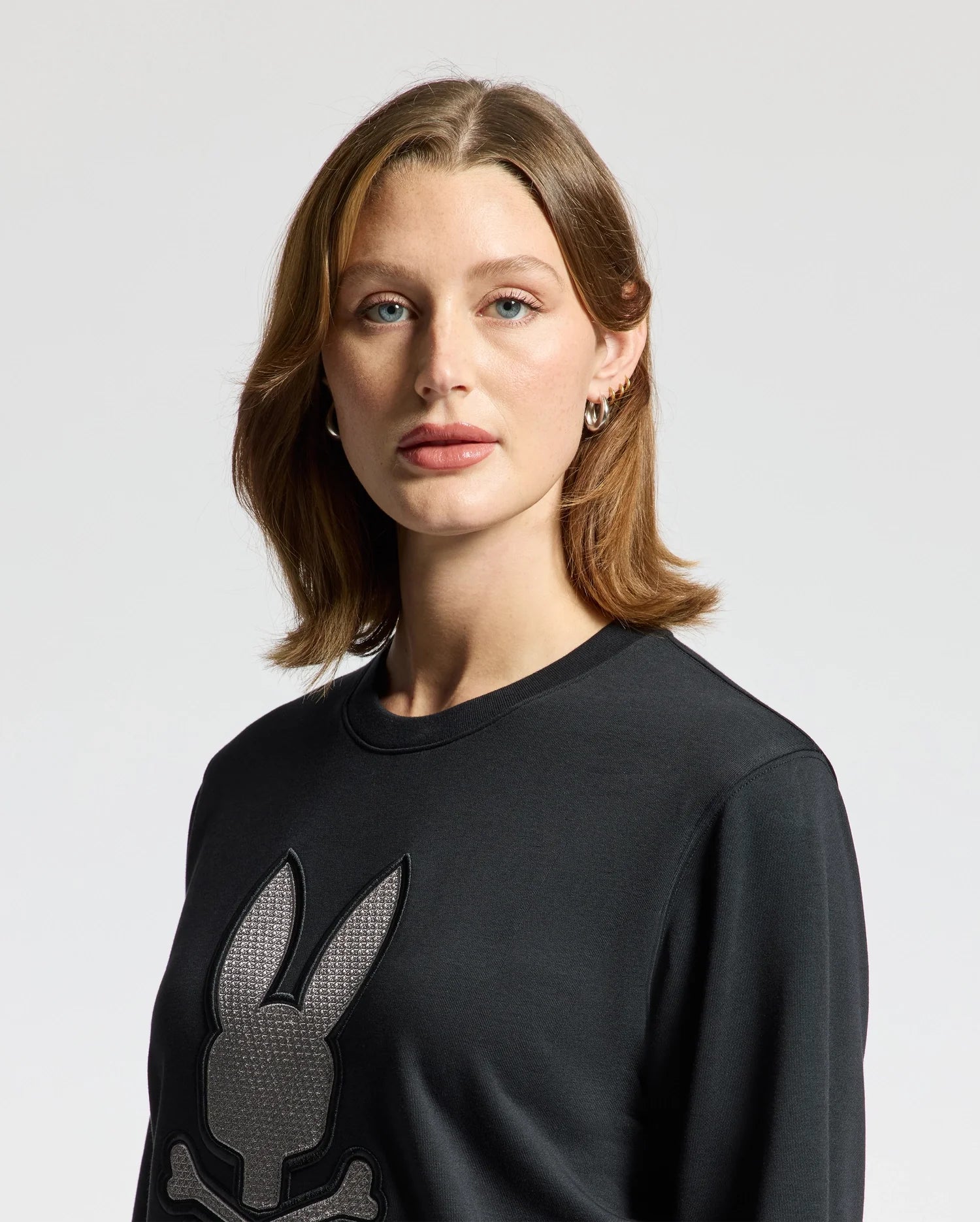 A woman with shoulder-length light brown hair wears the Psycho Bunny WOMENS ALEXANDER SWEATSHIRT - B2S156E200, a black cotton blend sweatshirt featuring an embroidered Lurex bunny design, standing against a neutral background.