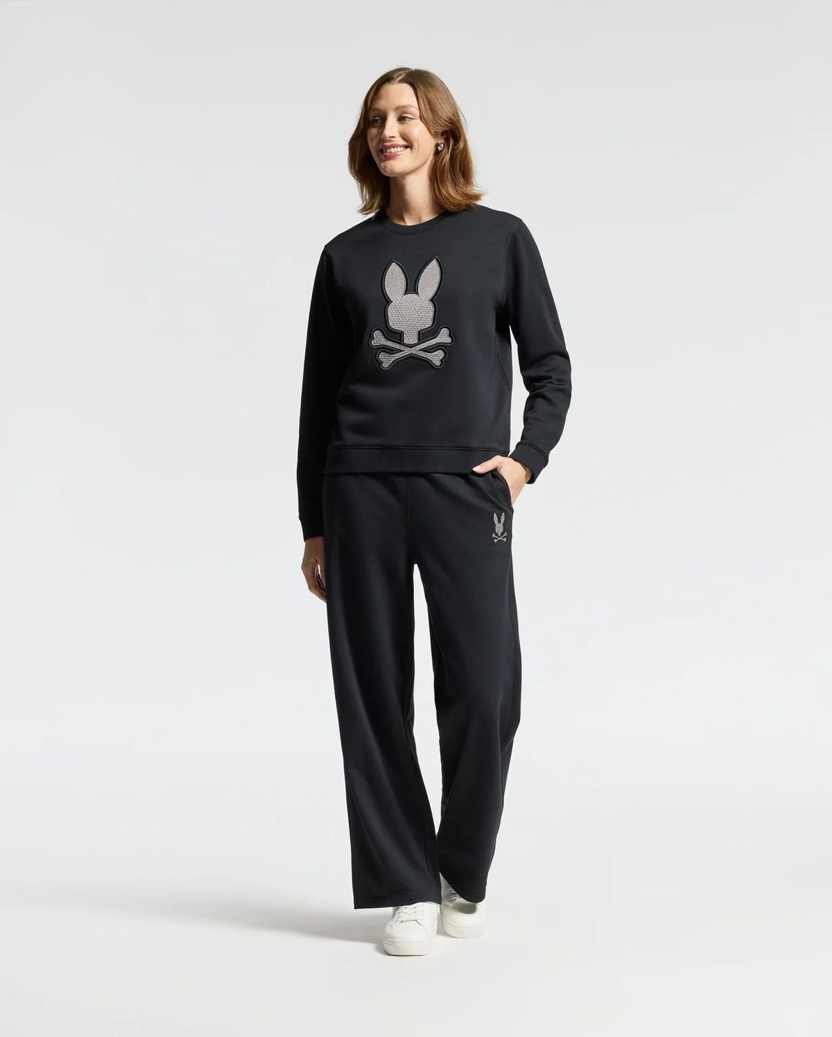 A person wearing the Psycho Bunny WOMENS ALEXANDER SWEATSHIRT - B2S156E200 in black, featuring an embroidered Lurex bunny and crossbones design, stands against a plain background. They are smiling with one hand casually in their pocket.