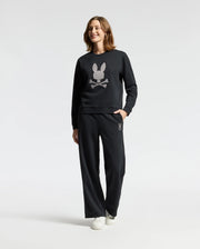 A person wearing the Psycho Bunny WOMENS ALEXANDER SWEATSHIRT - B2S156E200 in black, featuring an embroidered Lurex bunny and crossbones design, stands against a plain background. They are smiling with one hand casually in their pocket.