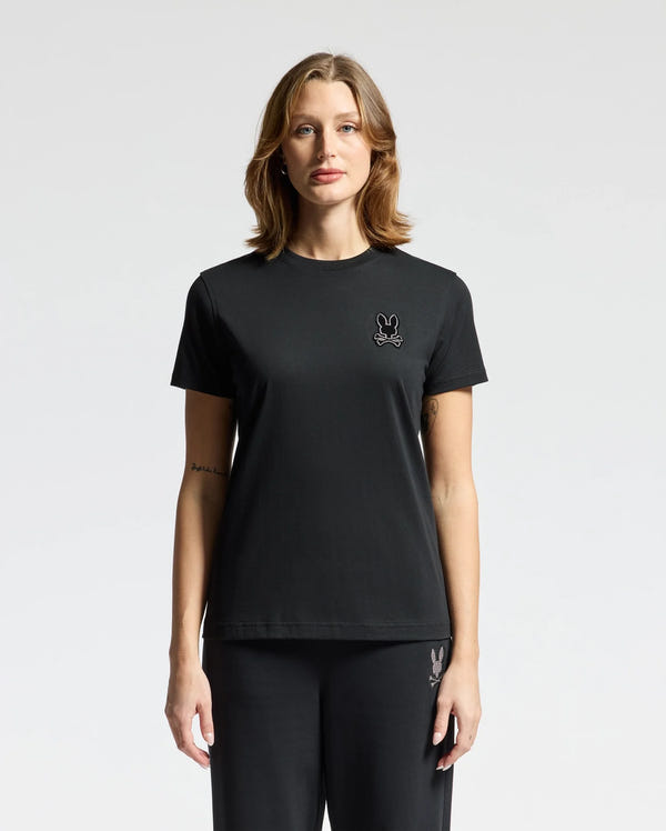 A woman wearing the Psycho Bunny WOMENS ALEXANDER GRAPHIC TEE (B2U164E200), crafted from soft Pima cotton, and black pants stands against a plain white background. The tee is adorned with a small rabbit logo featuring delicate lurex embroidery on the left chest.