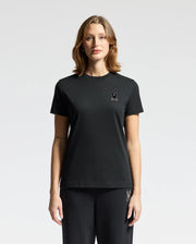 A woman wearing the Psycho Bunny WOMENS ALEXANDER GRAPHIC TEE (B2U164E200), crafted from soft Pima cotton, and black pants stands against a plain white background. The tee is adorned with a small rabbit logo featuring delicate lurex embroidery on the left chest.