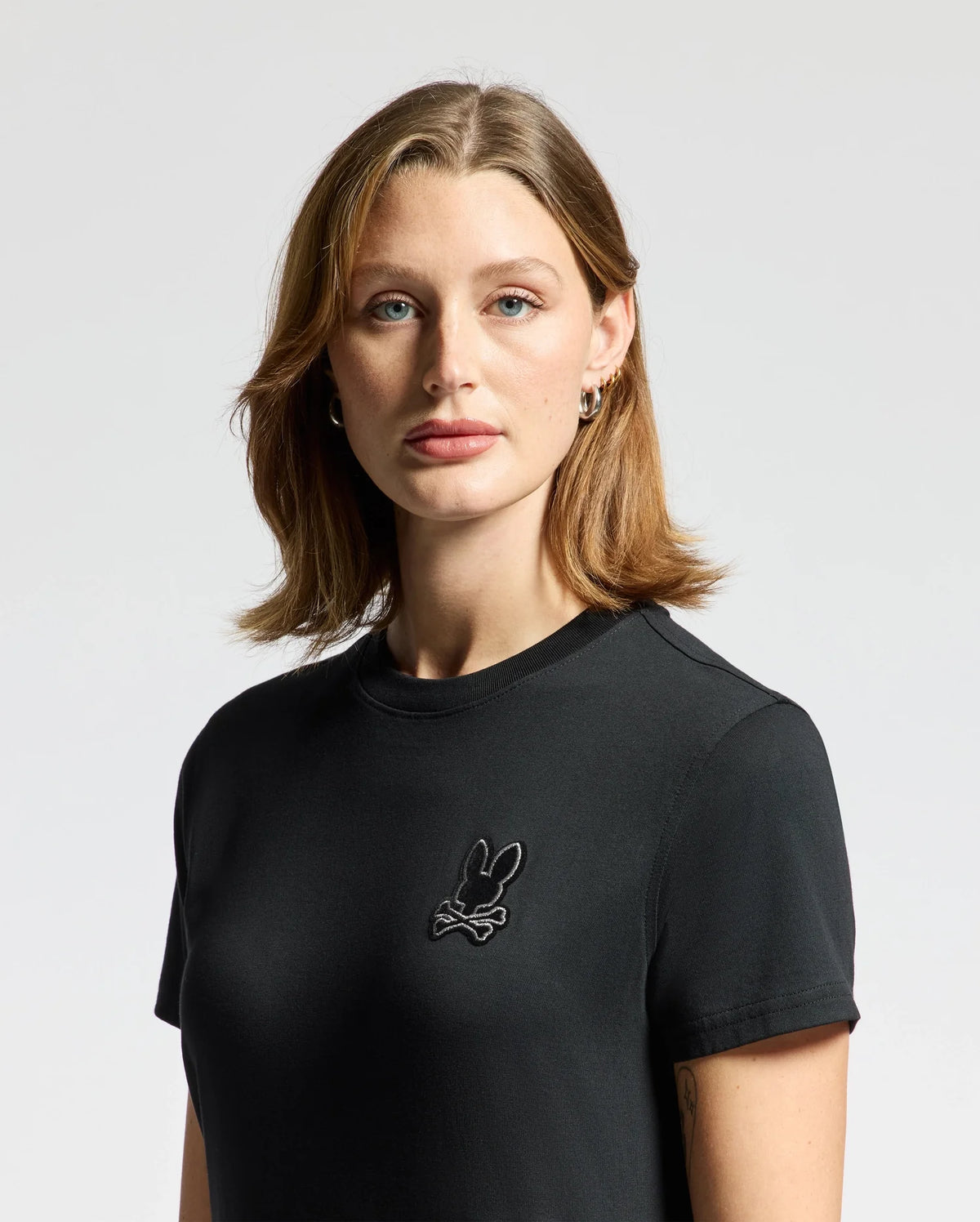 A person with shoulder-length brown hair is wearing a stylish Psycho Bunny Women's Alexander Graphic Tee (B2U164E200) in Pima Cotton, featuring a small Lurex Embroidery design on the chest. The background is plain and light-colored.