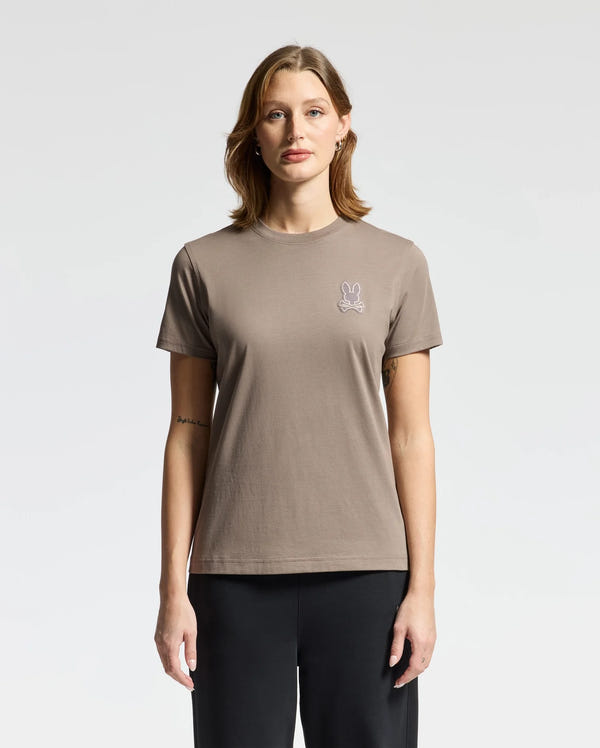 A person wearing the WOMENS ALEXANDER GRAPHIC TEE by Psycho Bunny, which is a taupe women's shirt featuring a small bunny logo possibly with lurex embroidery, stands against a plain white background. They have short brown hair and are paired with black pants.