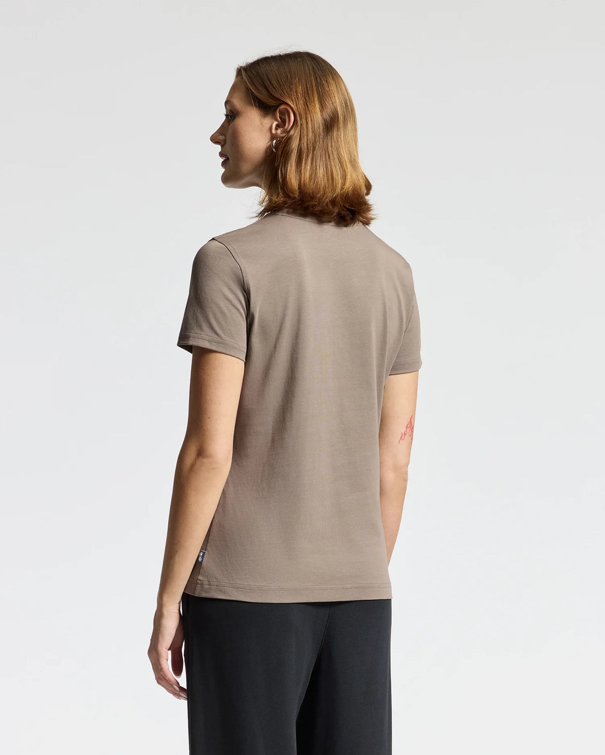 A person with shoulder-length hair, dressed in a taupe WOMENS ALEXANDER GRAPHIC TEE - B2U164E200 by Psycho Bunny, stands facing away from the camera. The soft Pima cotton fabric elegantly complements the simple white background.