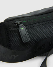 Close-up of the MENS CROSS BODY BAG - B6A181E200 by Psycho Bunny, crafted from black nylon and featuring a mesh pocket alongside a zipper pull adorned with a small bunny logo. The bag includes a strap with printed text adjacent to the pocket and boasts a subtle green accent near the zipper, combining style and functionality for everyday use.