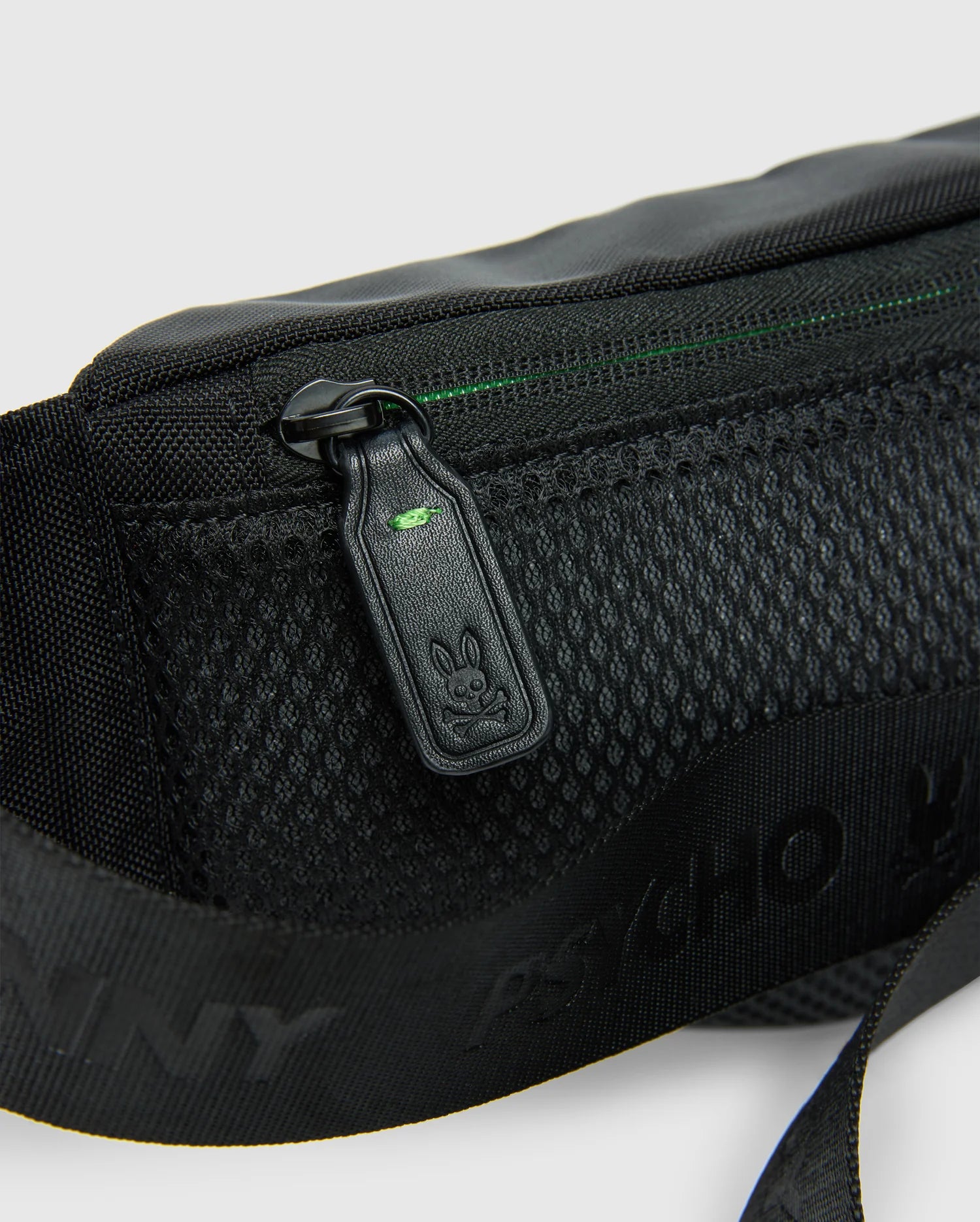 Close-up of the MENS CROSS BODY BAG - B6A181E200 by Psycho Bunny, crafted from black nylon and featuring a mesh pocket alongside a zipper pull adorned with a small bunny logo. The bag includes a strap with printed text adjacent to the pocket and boasts a subtle green accent near the zipper, combining style and functionality for everyday use.