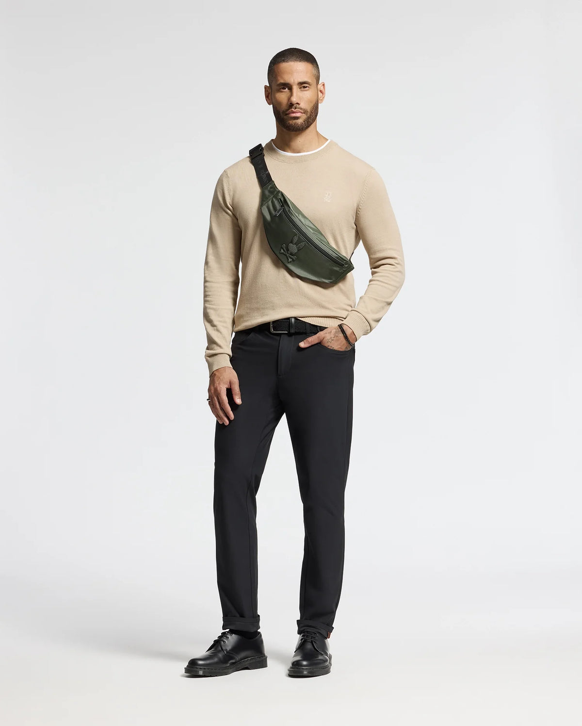 Against a white backdrop, a man wears a beige sweater, black pants, and shoes. A Psycho Bunny MENS CROSS BODY BAG - B6A181E200 in dark green is slung across his chest. With his right hand in his pocket, he exudes casual style with this chic accessory.
