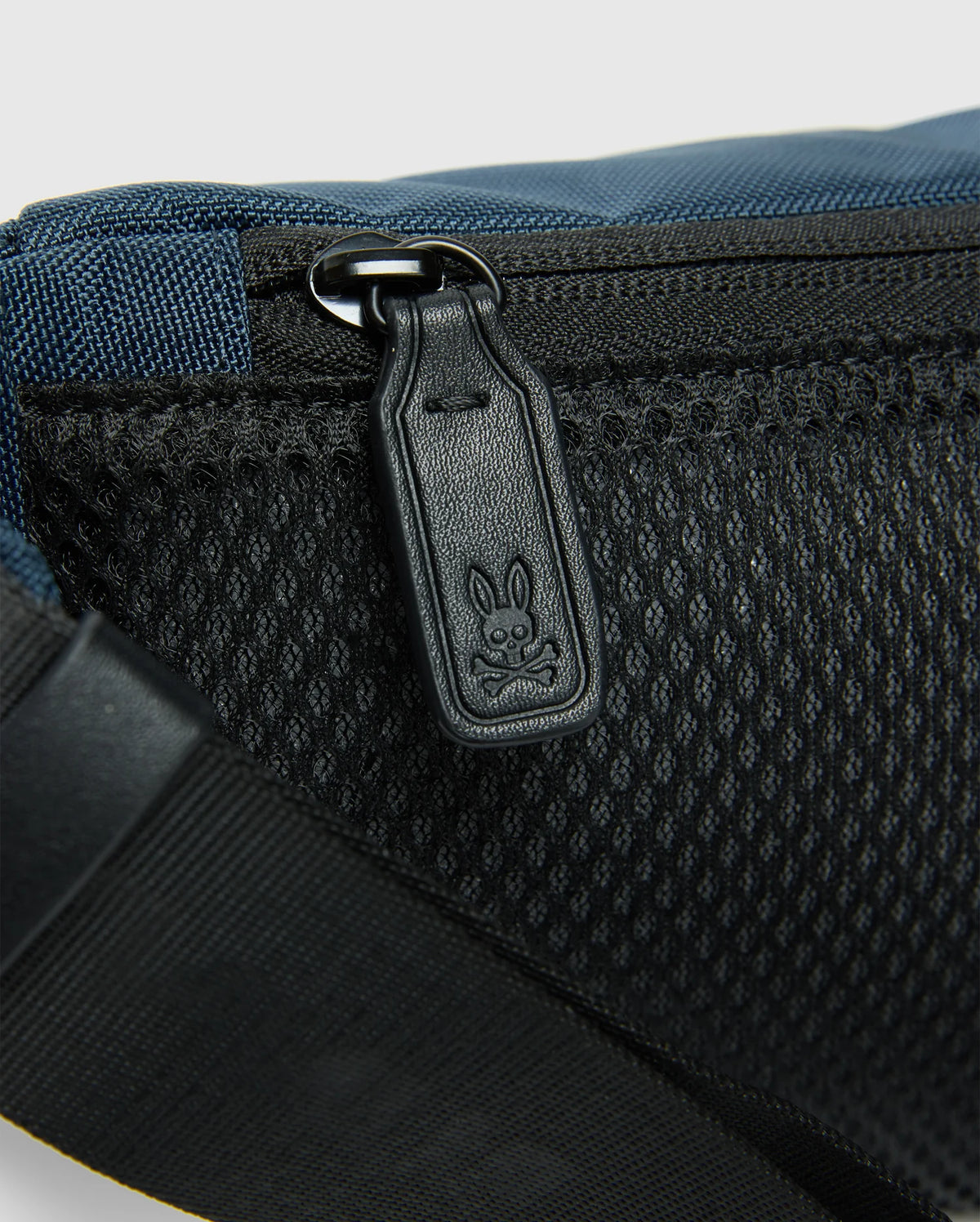 The MENS CROSS BODY BAG - B6A181E200 by Psycho Bunny highlights a detailed view of its blue and black design, emphasizing the black zipper pull engraved with a small rabbit motif. Crafted from durable nylon, this bag includes a mesh pocket and an adjustable strap for added versatility, making it ideal as a men's cross body accessory.