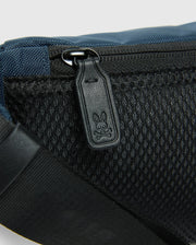 The MENS CROSS BODY BAG - B6A181E200 by Psycho Bunny highlights a detailed view of its blue and black design, emphasizing the black zipper pull engraved with a small rabbit motif. Crafted from durable nylon, this bag includes a mesh pocket and an adjustable strap for added versatility, making it ideal as a men's cross body accessory.