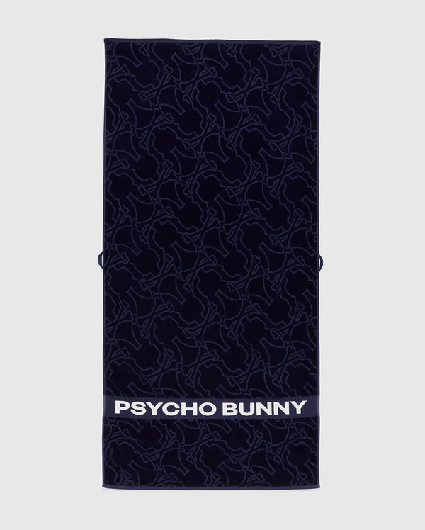 The Towel - B6A287F200 by Psycho Bunny is a rectangular dark towel made from premium terrycloth cotton, featuring an intricate bunny pattern. The bottom displays "PSYCHO BUNNY" in bold white letters, showcasing high-quality design.