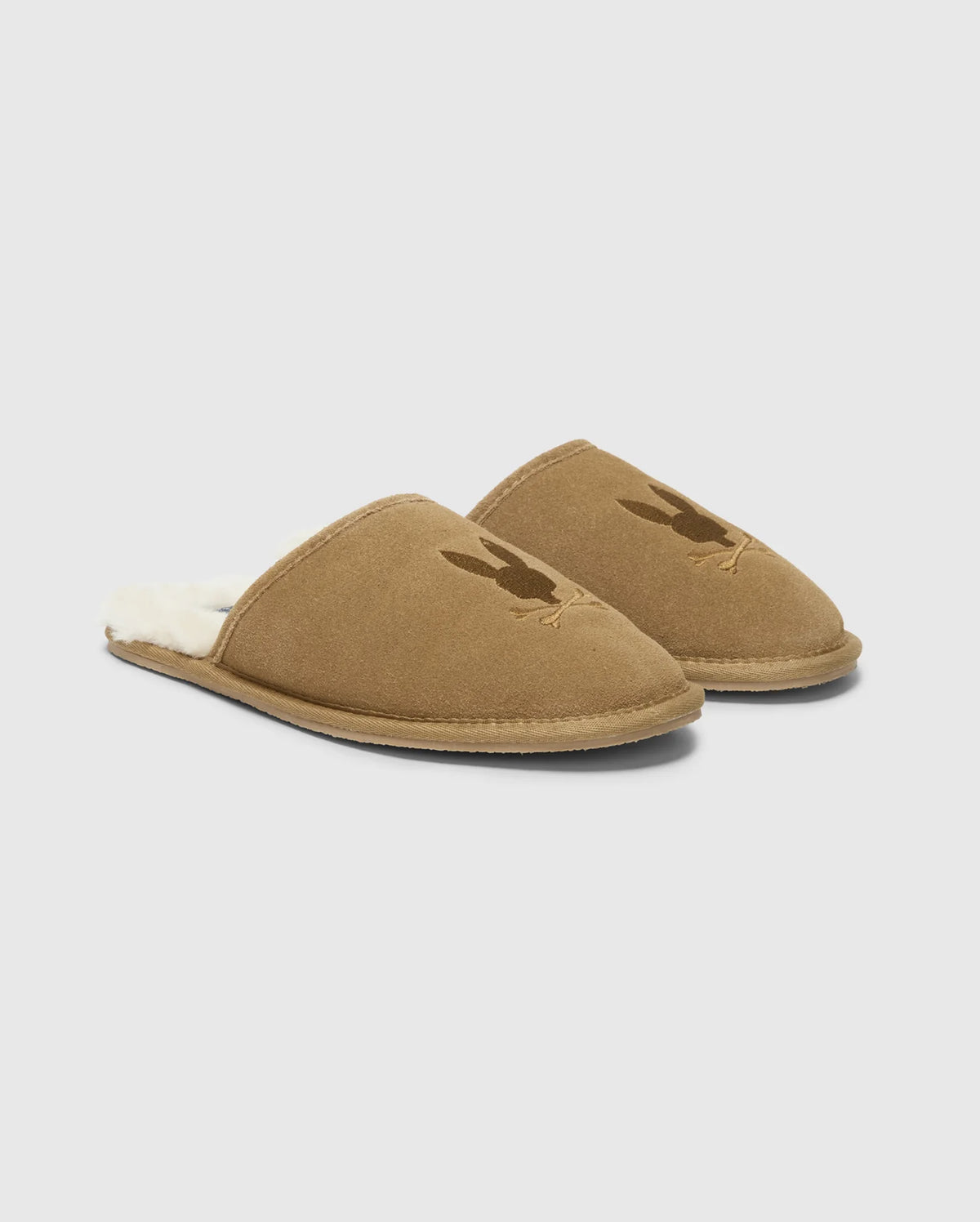 A pair of MENS SUEDE SLIPPERS by Psycho Bunny, crafted from cozy cow suede leather in brown with a soft, fuzzy interior. Each slipper is adorned with a subtle rabbit silhouette on the upper part, set against a clean white background.