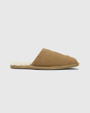 The MENS SUEDE SLIPPERS by Psycho Bunny, model B6A357E200, are crafted from brown cow suede leather and feature a plush off-white lining. These men's slippers have a flat sole and open back design, making them ideal for indoor comfort. The background is a subtle light gray.