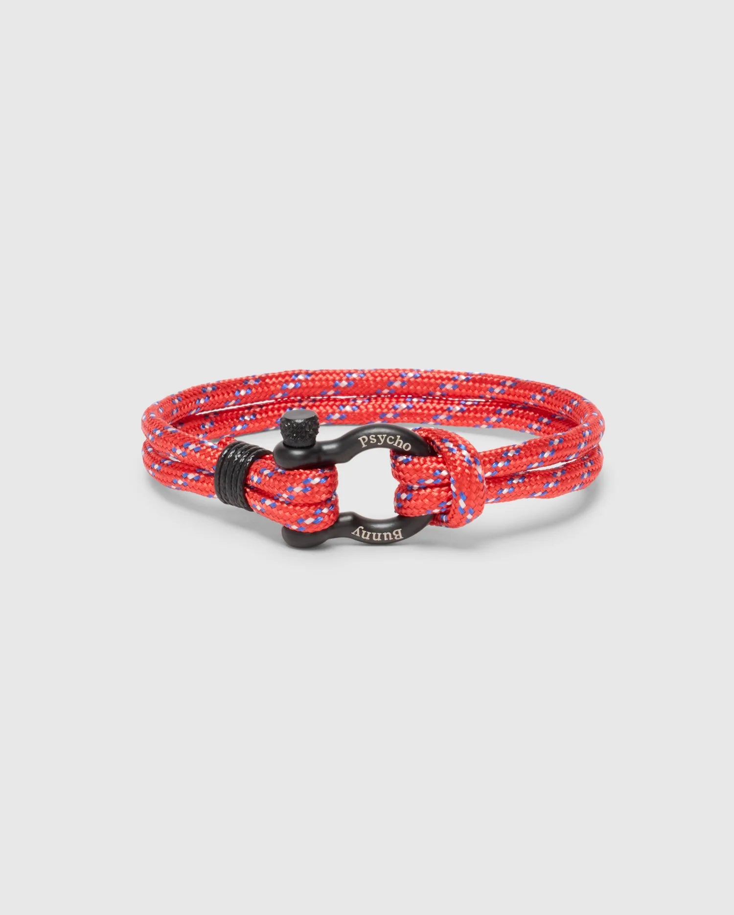 The Psycho Bunny Men's Rope Bracelet (B6A368E200) features a braided red and blue nylon rope with a black metal clasp engraved with "Psycho Bunny," adding sophistication. The elegant bracelet is neatly coiled on a light gray background.