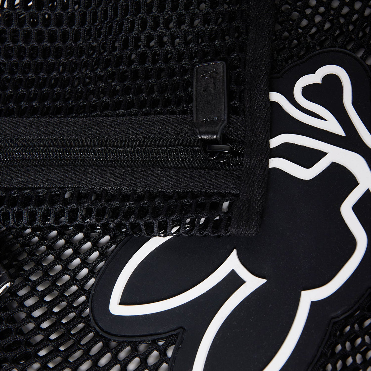 Close-up of a black mesh tote material with a zipper. The zipper pull tab is visible, alongside a black and white stylized graphic of a figure with outstretched arms and legs peeking through the mesh. The Psycho Bunny MENS MESH TOTE BAG - B6A388B200 features detachable zip pouch for added convenience.