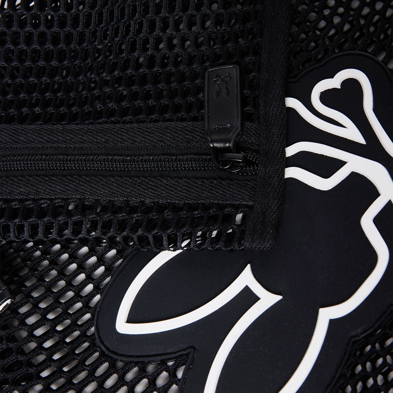 Close-up of a black mesh tote material with a zipper. The zipper pull tab is visible, alongside a black and white stylized graphic of a figure with outstretched arms and legs peeking through the mesh. The Psycho Bunny MENS MESH TOTE BAG - B6A388B200 features detachable zip pouch for added convenience.