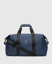 The MENS DUFFLE BAG - B6A472F200 by Psycho Bunny is a sleek blue travel accessory with black straps and handles, featuring their raised bunny with crossbones logo. It includes "Psycho Bunny" on the quality straps and offers a detachable shoulder strap for added convenience.