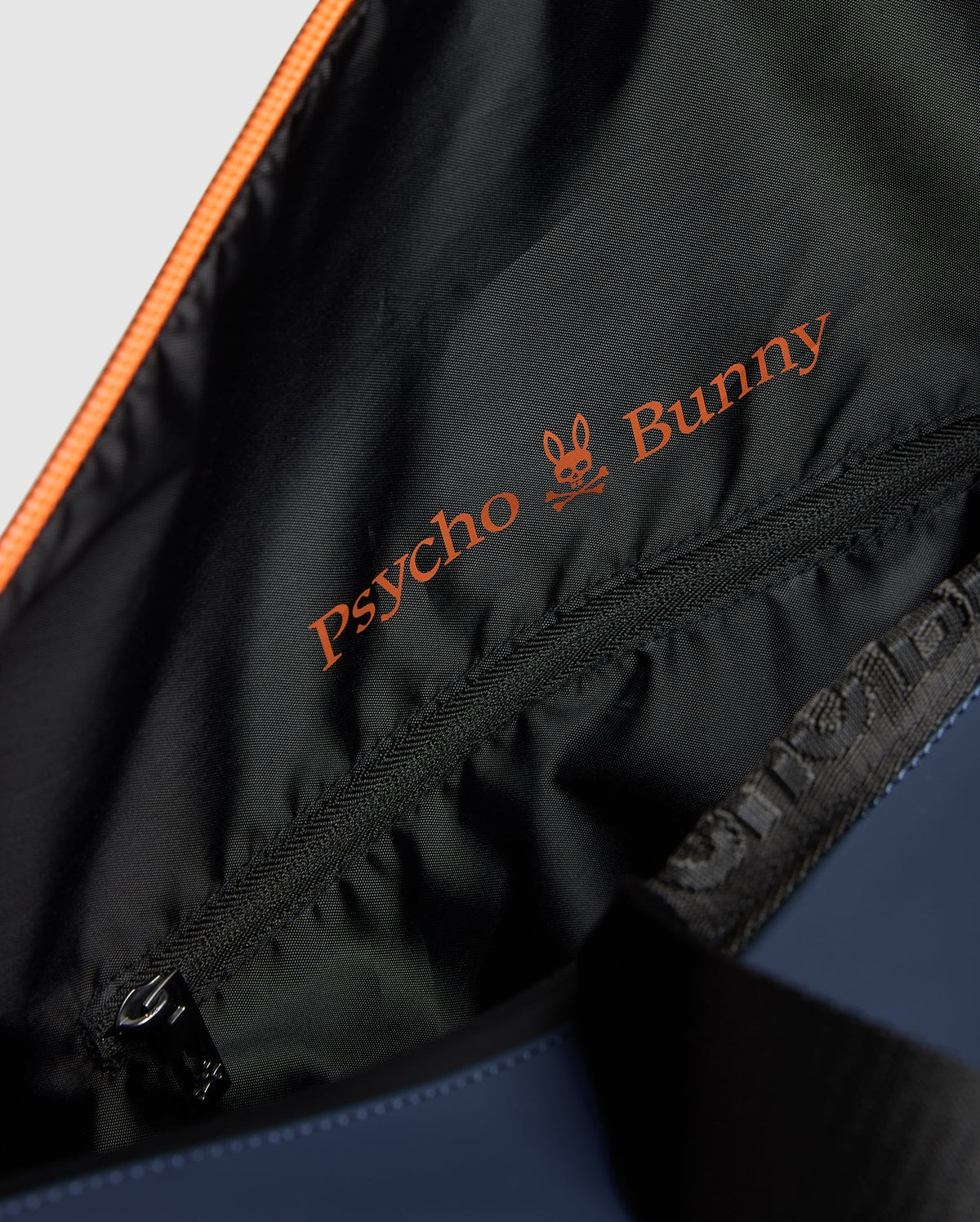 A close-up of the MENS DUFFLE BAG - B6A472F200 reveals the vibrant orange embroidered "Psycho Bunny" logo on its black lining, adorned with a stylish bunny skull design. A gleaming zipper adds a touch of quality craftsmanship.