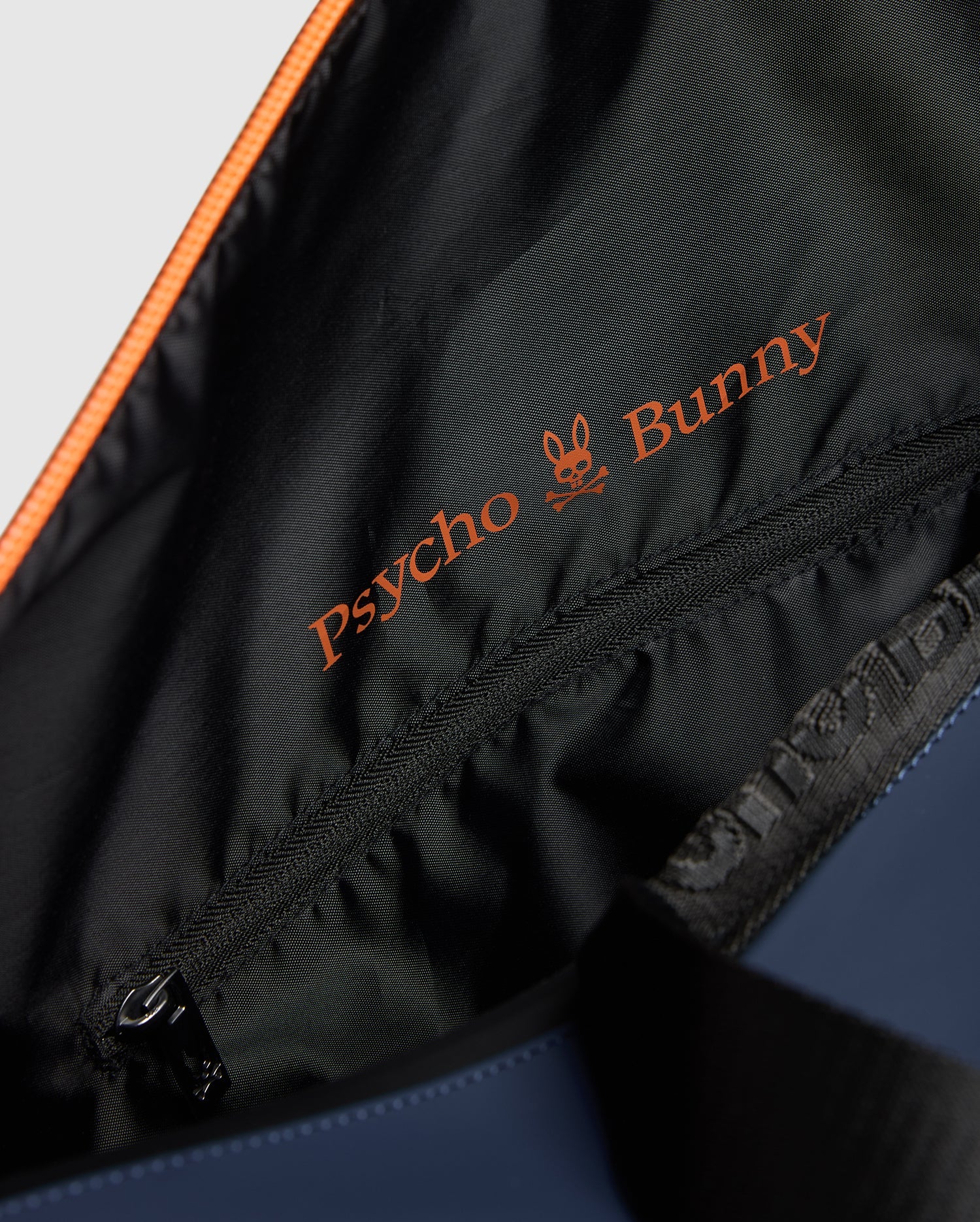 A close-up of the MENS DUFFLE BAG - B6A472F200 reveals the vibrant orange embroidered "Psycho Bunny" logo on its black lining, adorned with a stylish bunny skull design. A gleaming zipper adds a touch of quality craftsmanship.
