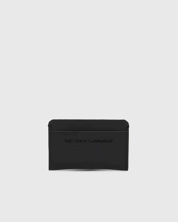 The Psycho Bunny MENS RUBBERIZED CARD CASE - B6A473F200, shown in black against a white background, offers peace of mind with its sleek design. Embossed with "THE BOLD STANDARD," this minimalist card case keeps your essentials safe.