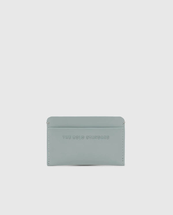 The Psycho Bunny MENS RUBBERIZED CARD CASE - B6A473F200 features "THE BOLD STANDARD" embossed on its light gray, minimalist leather design. Offering security and elegance against a white background, this piece is both stylish and functional.