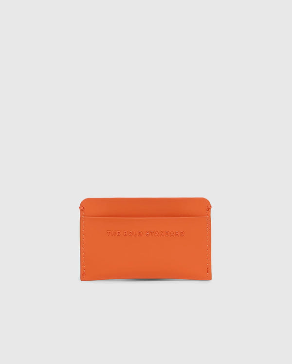 The Psycho Bunny MENS RUBBERIZED CARD CASE - B6A473F200 is an orange leather card case embossed with "THE BOLD STANDARD." Its minimalist design features rounded corners and a slim profile, perfect for stylishly storing your essentials against a light grey background.