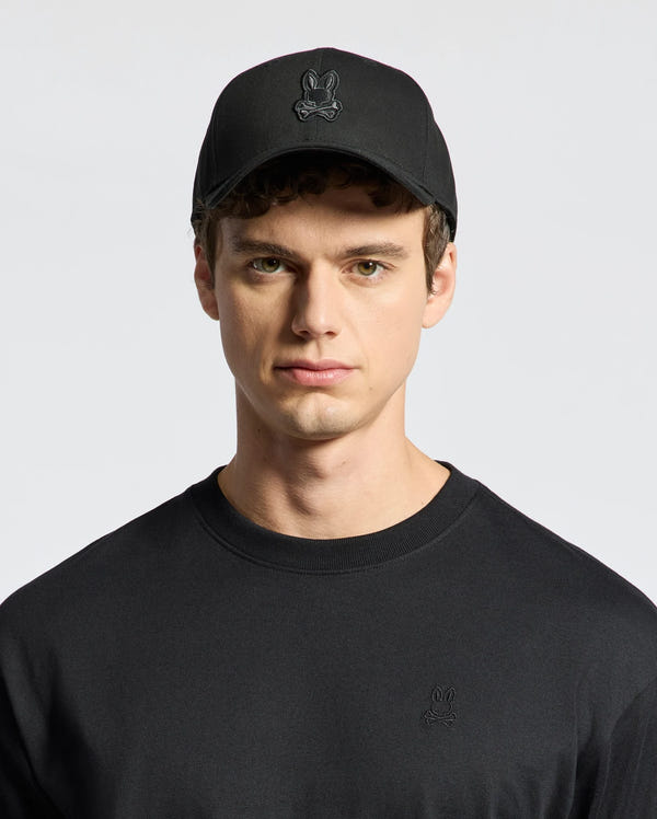 Dressed in a Psycho Bunny MENS NEW CASTLE GRAPHIC BASEBALL CAP - B6A547F200, featuring black embroidery and an adjustable back strap, paired with a matching cotton twill shirt against a plain white background.