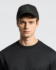 Dressed in a Psycho Bunny MENS NEW CASTLE GRAPHIC BASEBALL CAP - B6A547F200, featuring black embroidery and an adjustable back strap, paired with a matching cotton twill shirt against a plain white background.