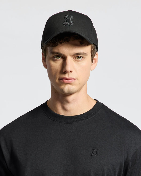 Dressed in a Psycho Bunny MENS NEW CASTLE GRAPHIC BASEBALL CAP - B6A547F200, featuring black embroidery and an adjustable back strap, paired with a matching cotton twill shirt against a plain white background.