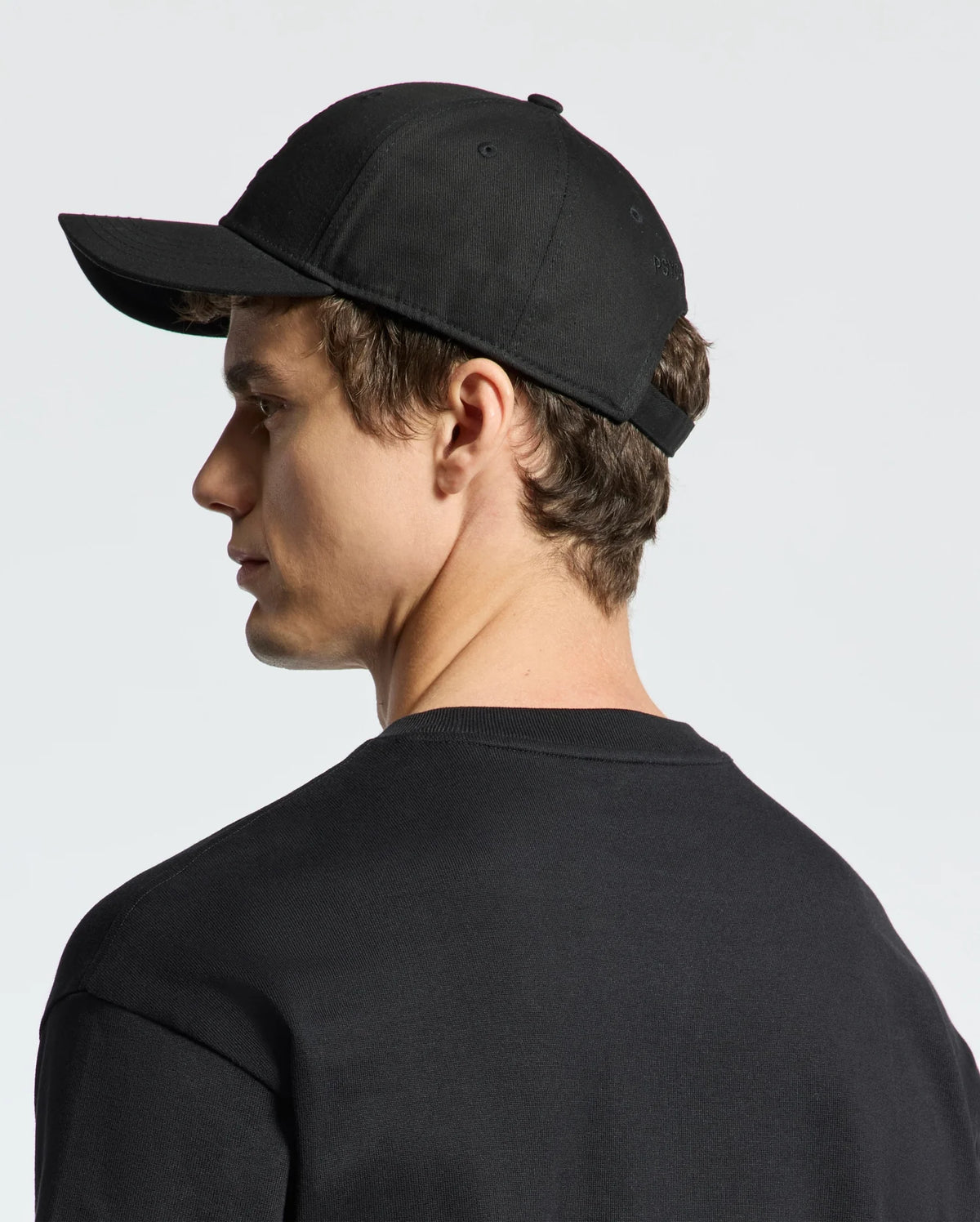 A person with short brown hair is wearing a Psycho Bunny MENS NEW CASTLE GRAPHIC BASEBALL CAP - B6A547F200. The black cap, paired with a black shirt, has an embroidered design, adjustable back strap, and cotton twill texture. They're facing left against a plain light background.