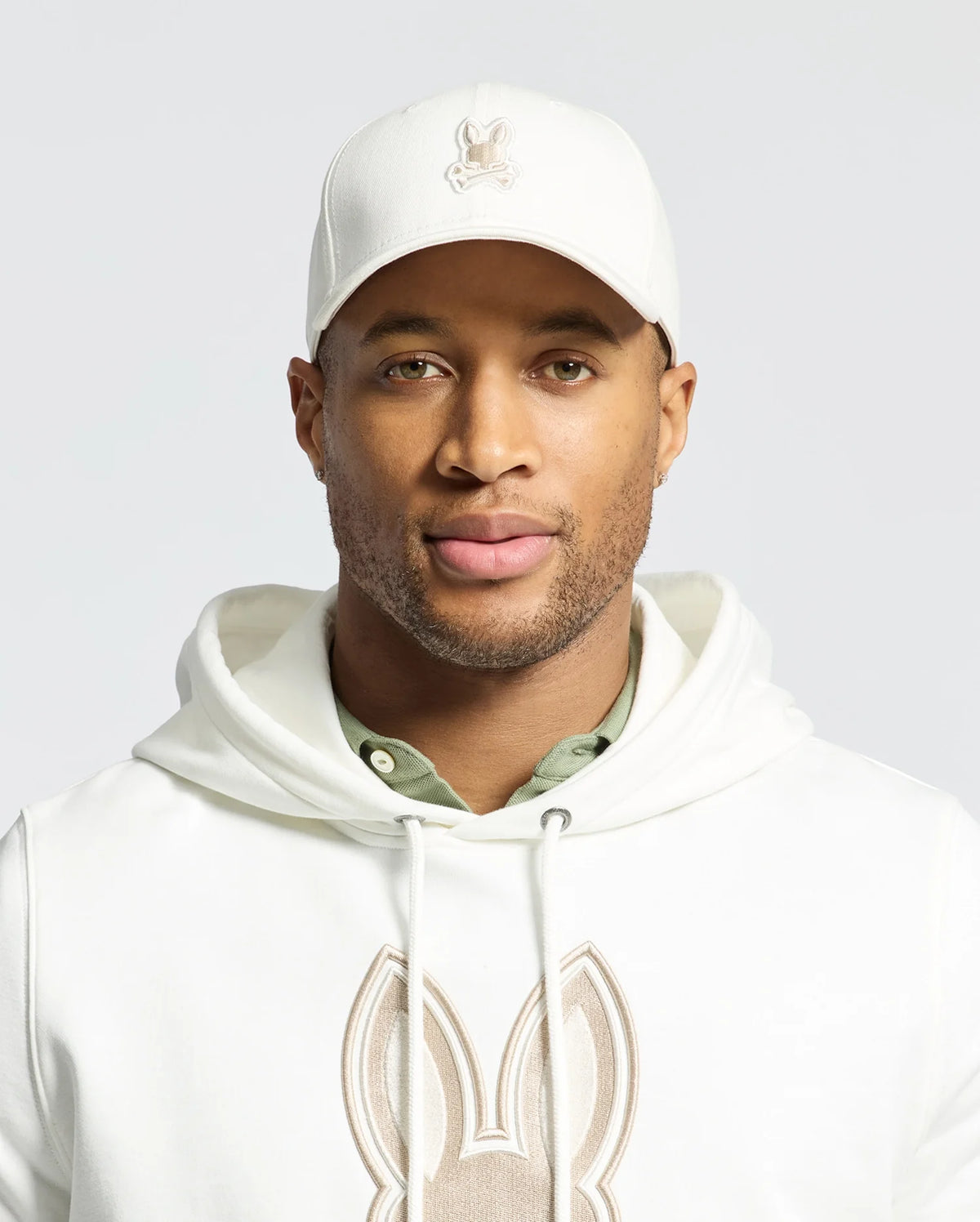 A man proudly wears the Psycho Bunny MENS NEW CASTLE GRAPHIC BASEBALL CAP in white cotton twill with an embroidered bunny logo, paired with a matching hoodie. A green shirt subtly emerges, embodying quiet luxury as he gazes confidently at the camera against a plain background.