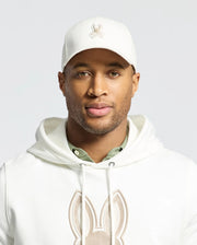 A man proudly wears the Psycho Bunny MENS NEW CASTLE GRAPHIC BASEBALL CAP in white cotton twill with an embroidered bunny logo, paired with a matching hoodie. A green shirt subtly emerges, embodying quiet luxury as he gazes confidently at the camera against a plain background.