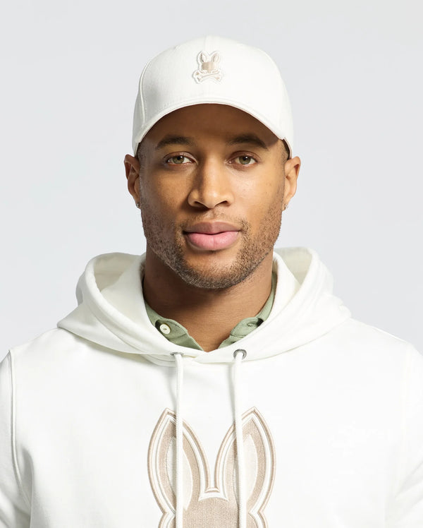 A man proudly wears the Psycho Bunny MENS NEW CASTLE GRAPHIC BASEBALL CAP in white cotton twill with an embroidered bunny logo, paired with a matching hoodie. A green shirt subtly emerges, embodying quiet luxury as he gazes confidently at the camera against a plain background.